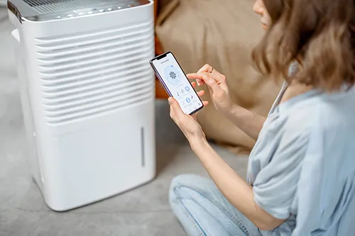 A woman sitting on the floor and using a smartphone app to adjust settings on a modern white home dehumidifier.