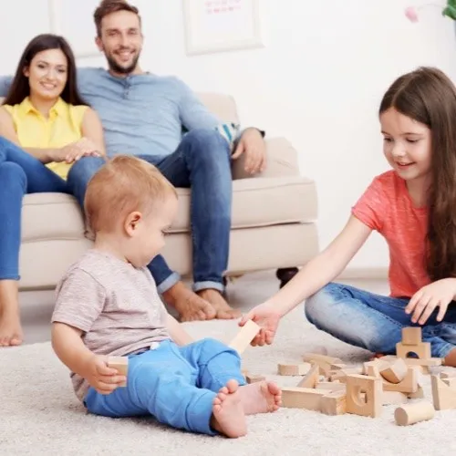 A toddler and a young girl playing with wooden blocks on a plush rug while their parents watch from a nearby sofa.
