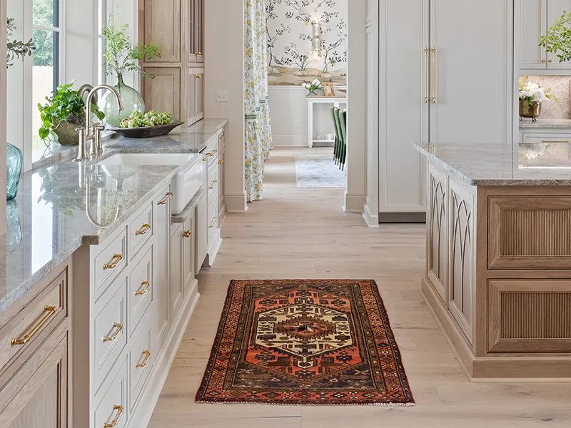 A long, modern kitchen with white cabinets, light wood floors, grey granite countertops, and a traditional, patterned Persian runner rug in the center aisle.