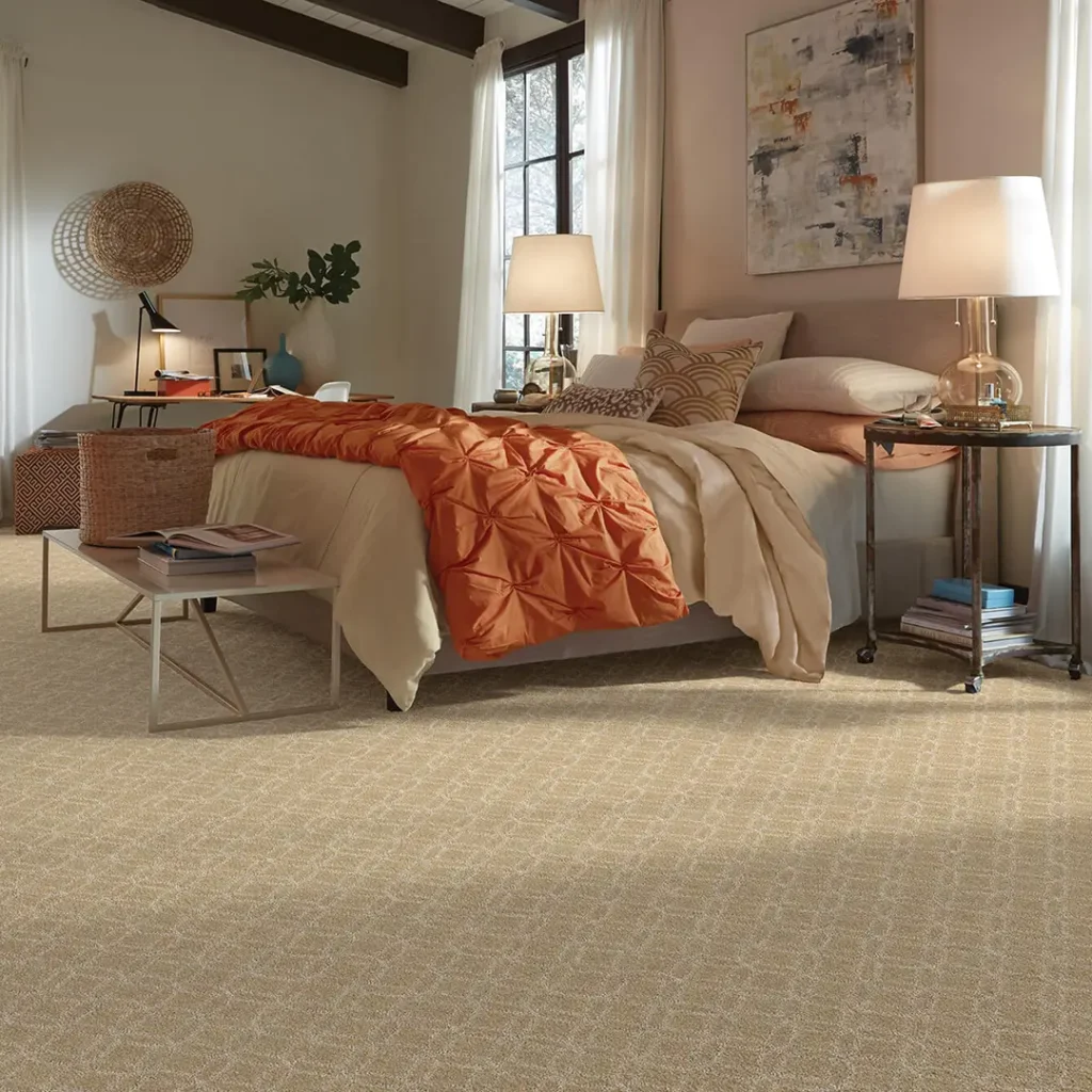 Patterned beige wall-to-wall carpet in a cozy bedroom featuring a bed with a bright orange quilted coverlet and warm bedside lighting.