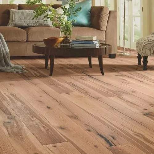 A low-angle view of rustic, medium-toned wide-plank hardwood flooring with prominent natural wood grain and knots, set in a cozy living room with a brown sofa and a round coffee table.
