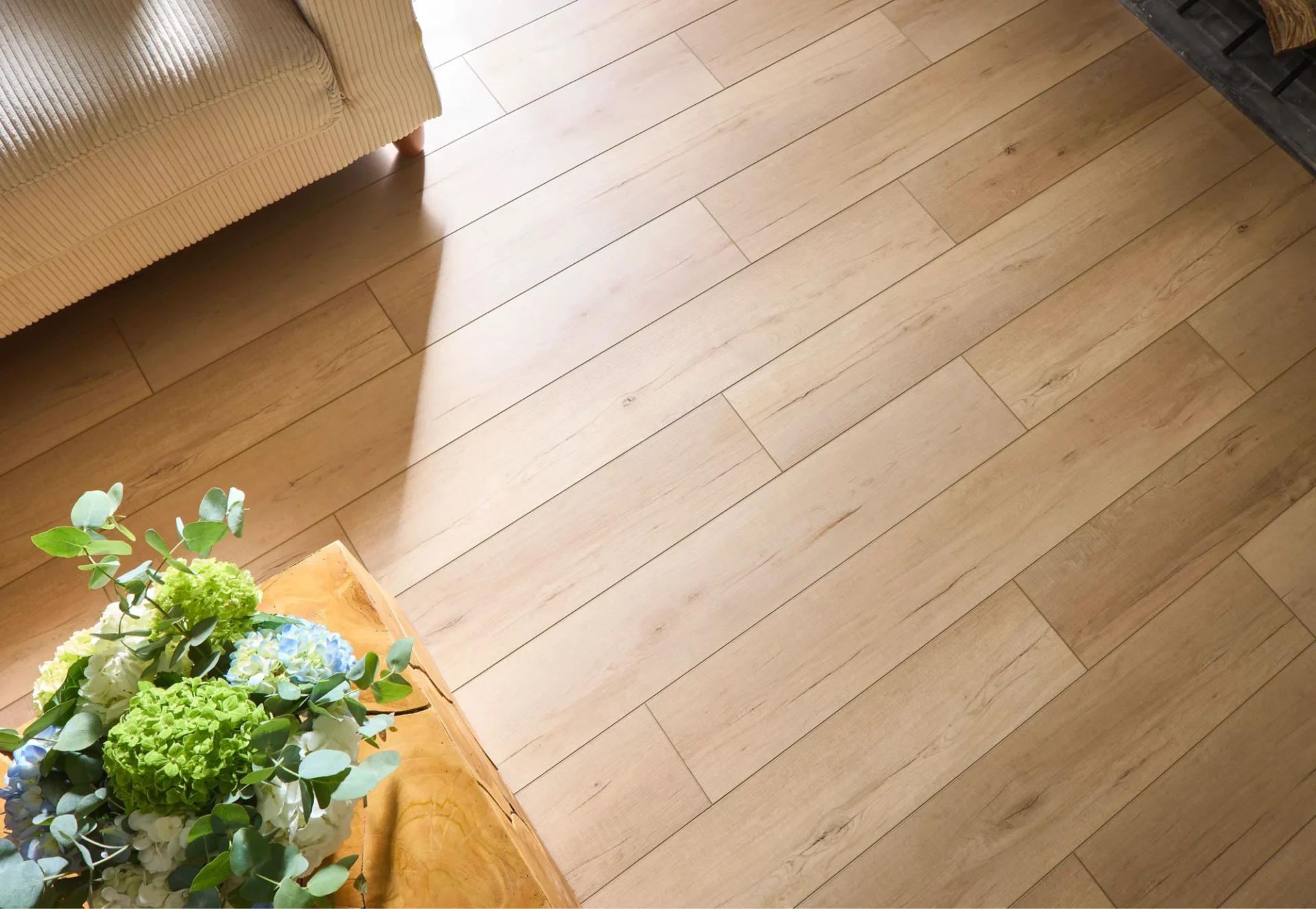 High-angle view of smooth, light-toned hardwood planks illuminated by bright sunlight next to a vibrant floral arrangement.