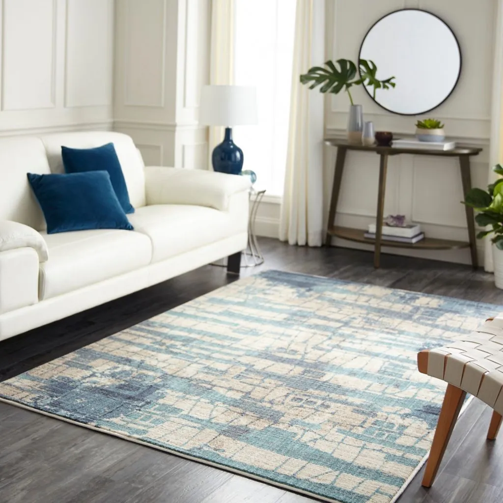 A bright living room showcasing a contemporary abstract area rug in shades of blue, teal, and cream beneath a white sofa with deep blue pillows.