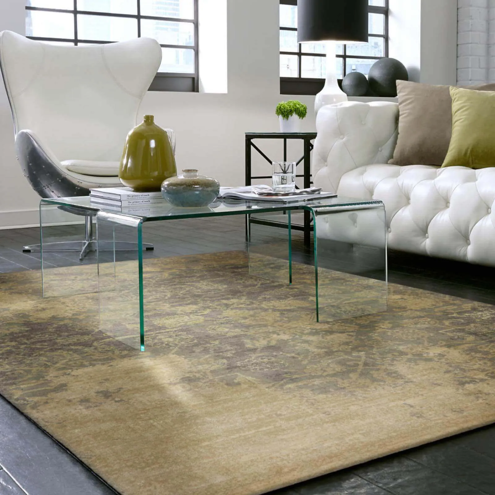 A modern living space with a muted olive and gold distressed area rug, a clear glass coffee table, and a tufted white leather sofa.