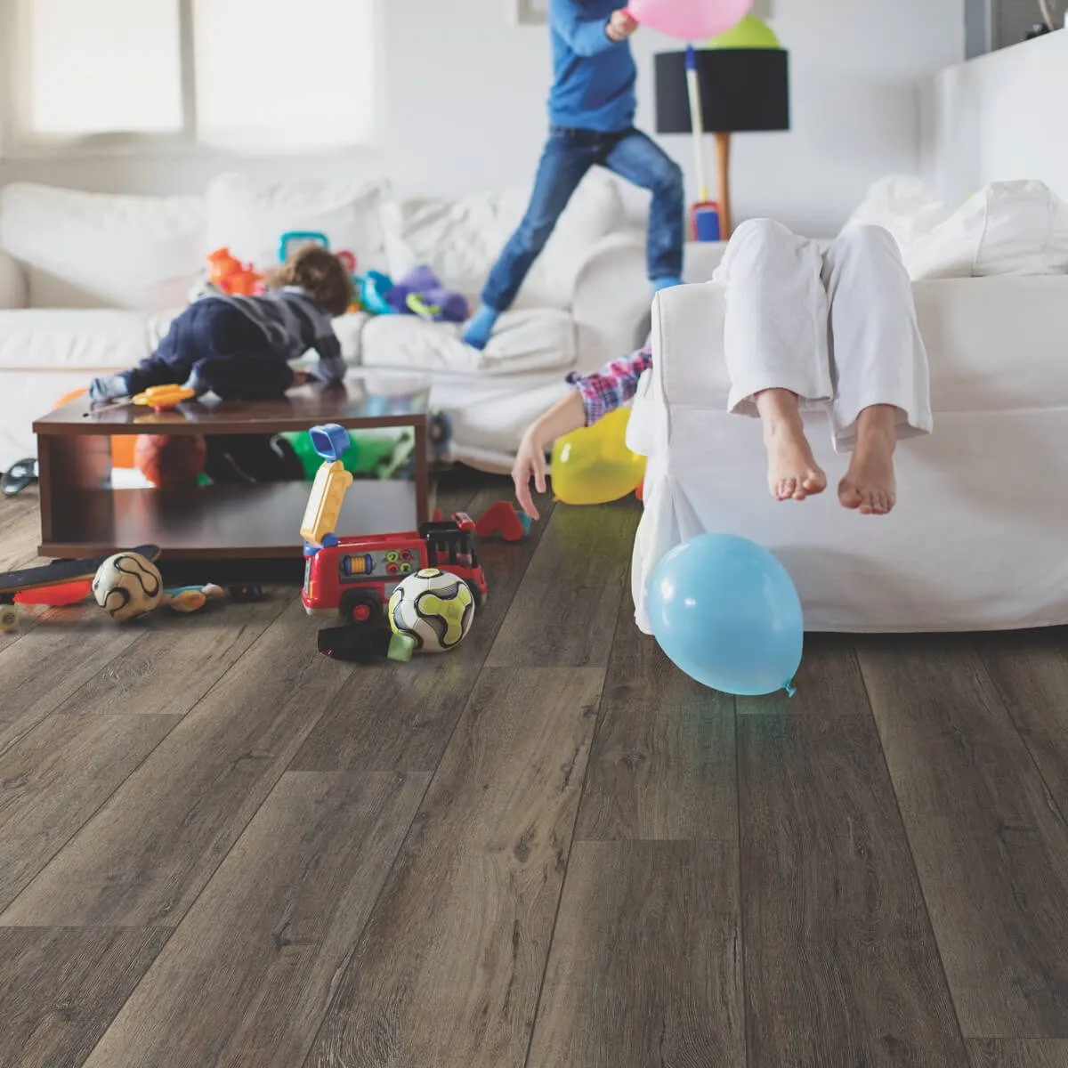 Dark wood-plank vinyl flooring in a busy, messy living room with kids playing, scattered toys, and colorful balloons.